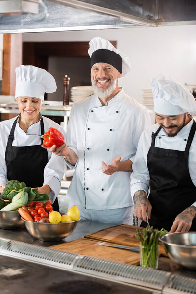 Happy chef holding bell pepper near interracial colleagues and vegetables in kitchen 