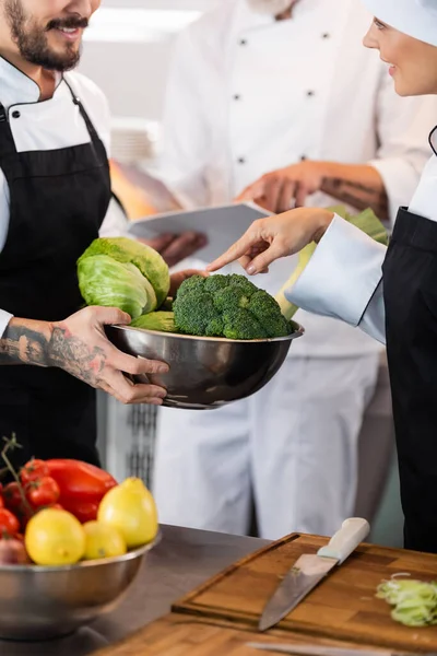 Usmívající Kuchař Ukazuje Zeleninu Misce Poblíž Kolegy Kuchyni — Stock fotografie