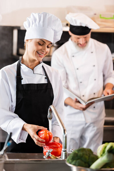 Smiling chef washing vegetables near colleague with cookbook in kitchen 