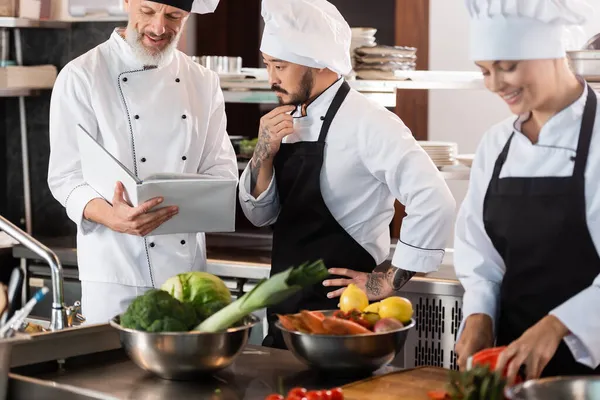 Sonriente Chef Celebración Libro Cocina Cerca Asiático Colega Verduras Cocina — Foto de Stock