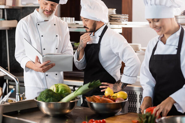 Smiling chef holding cookbook near asian colleague and vegetables in kitchen 