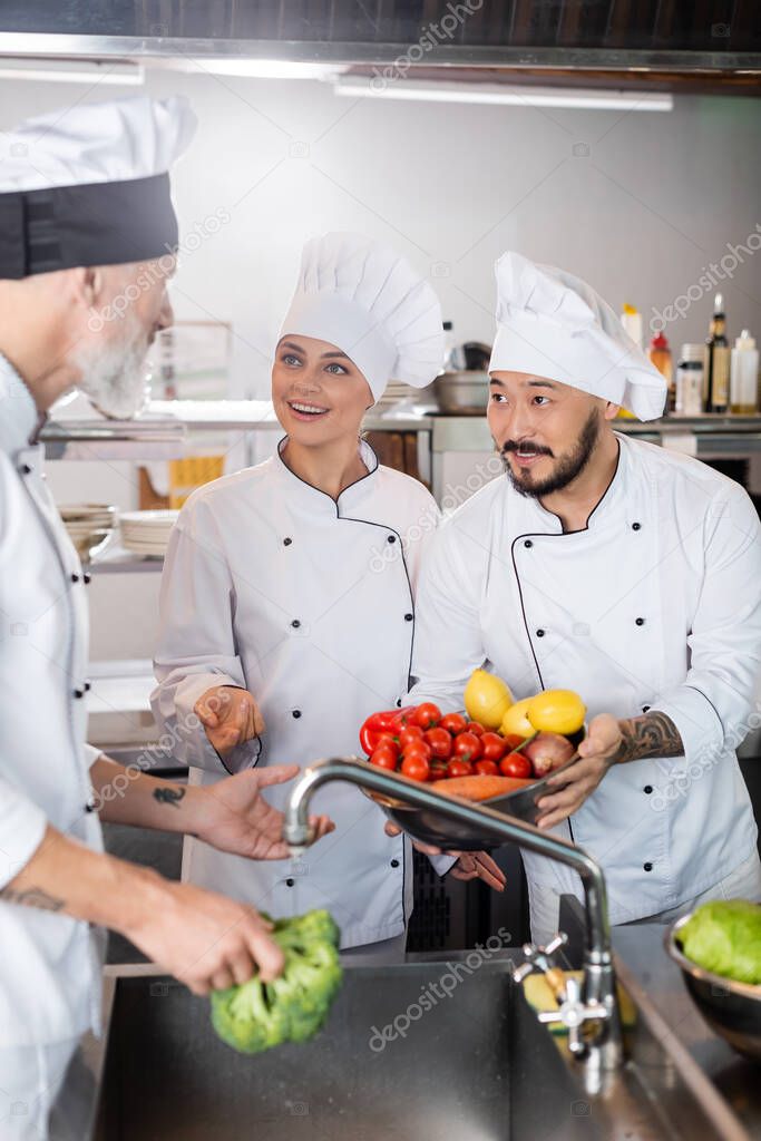 Multiethnic chefs with vegetables talking to colleague near sink in kitchen 