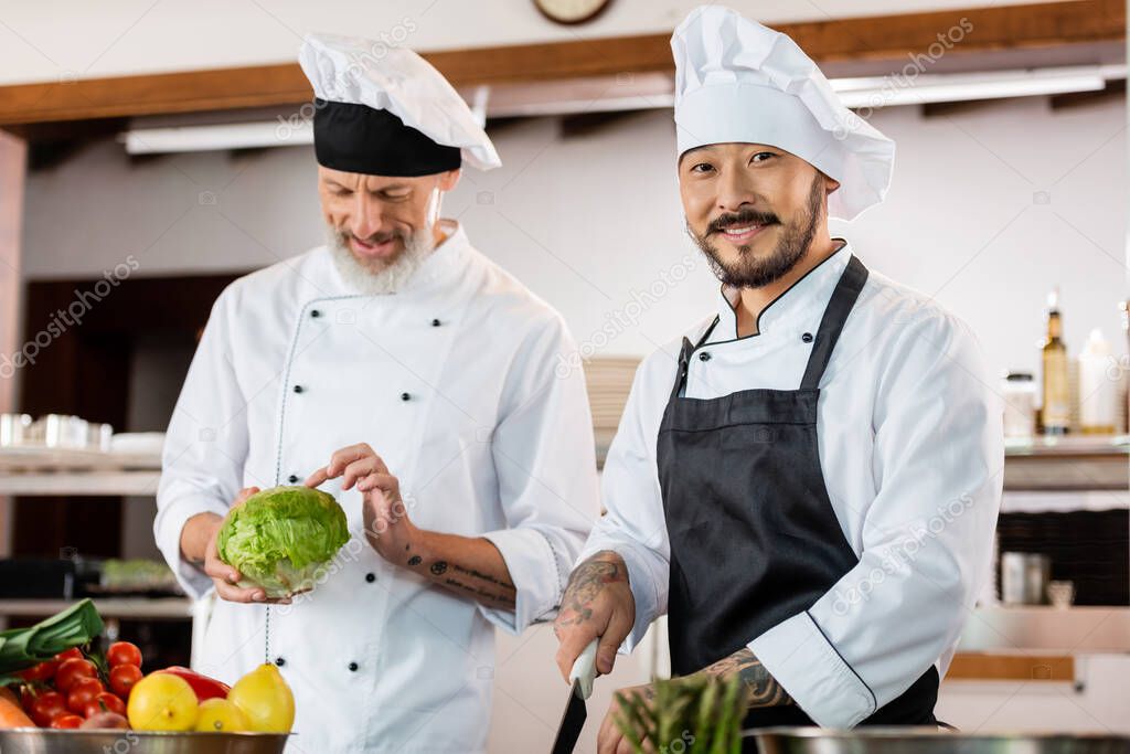 Asian chef with knife smiling at camera near blurred colleague and vegetables in kitchen 