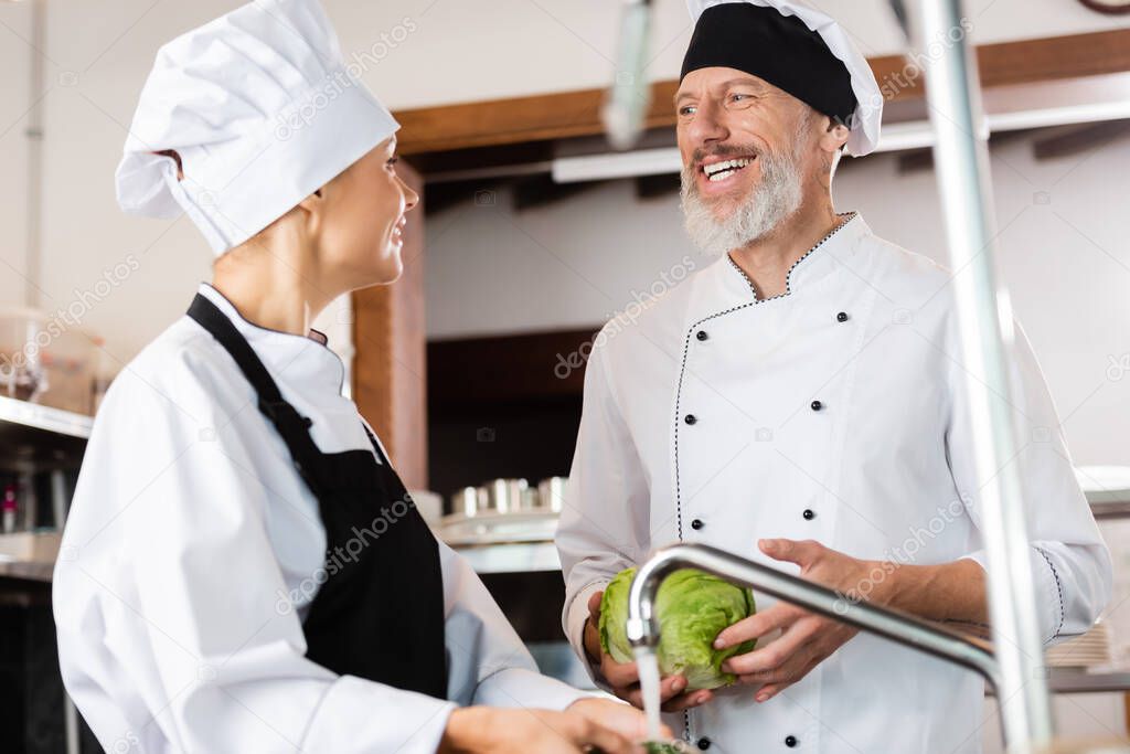 Positive chef holding cabbage near colleague and faucet in kitchen 