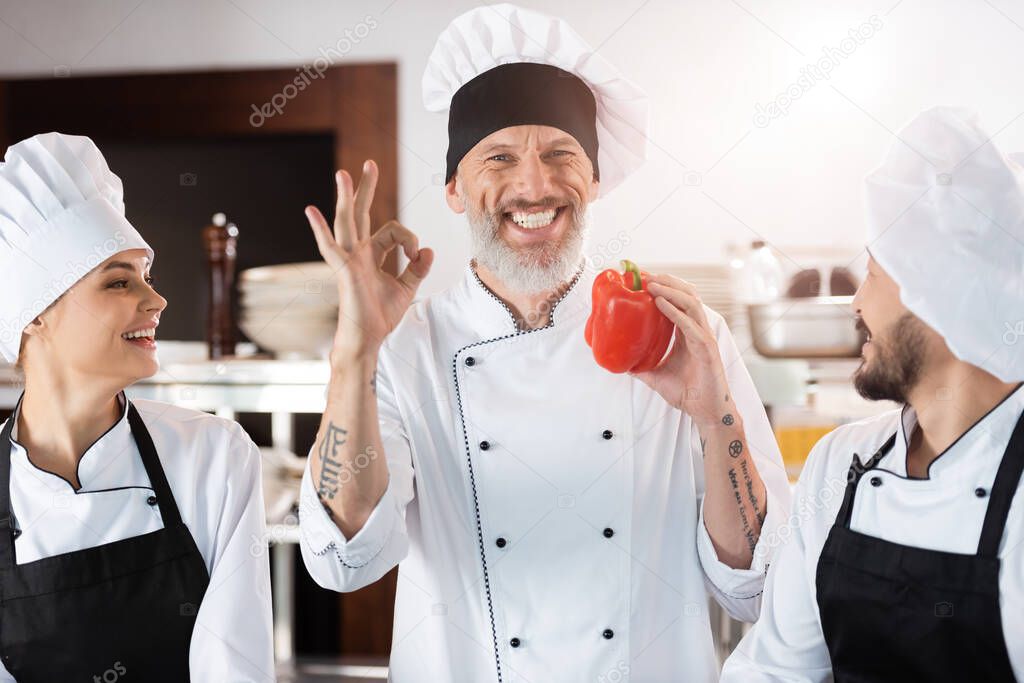 Smiling chef showing ok and holding bell pepper near multiethnic colleagues in kitchen 