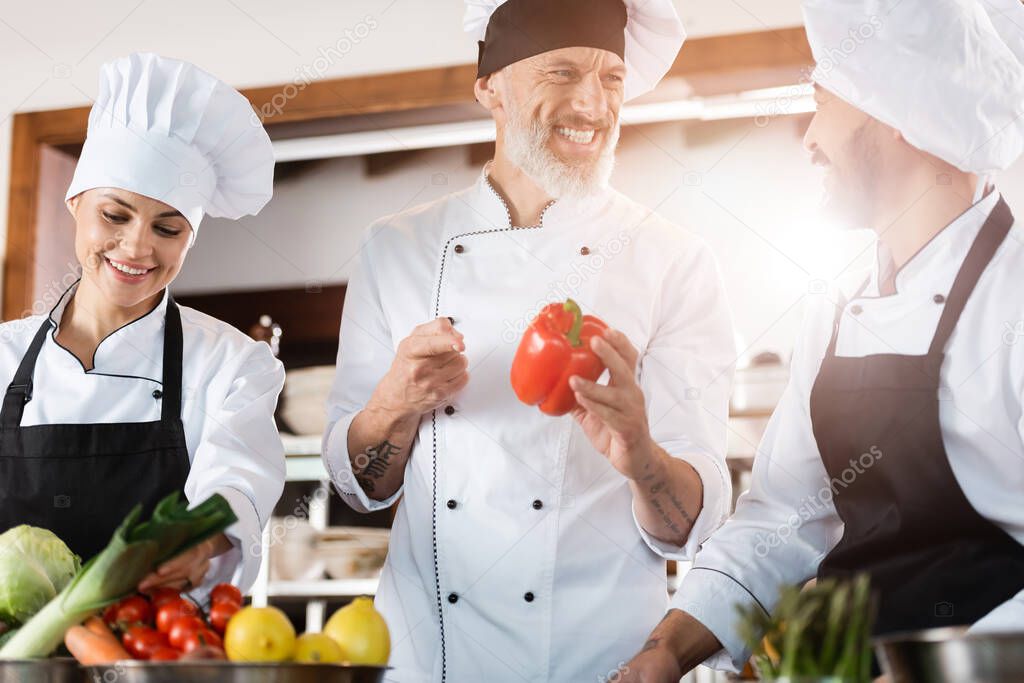 Chef pointing at bell pepper near smiling asian colleague in restaurant kitchen 
