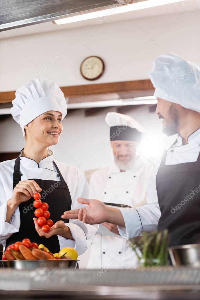 Smiling asian chef pointing at cherry tomatoes near colleague and ripe vegetables in kitchen 