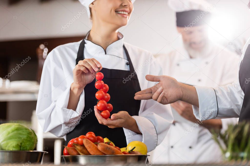 Chef pointing at cherry tomatoes near smiling colleague in restaurant kitchen 