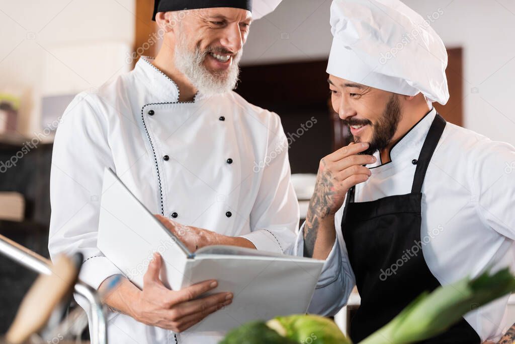 Cheerful interracial chefs working with cookbook in kitchen 