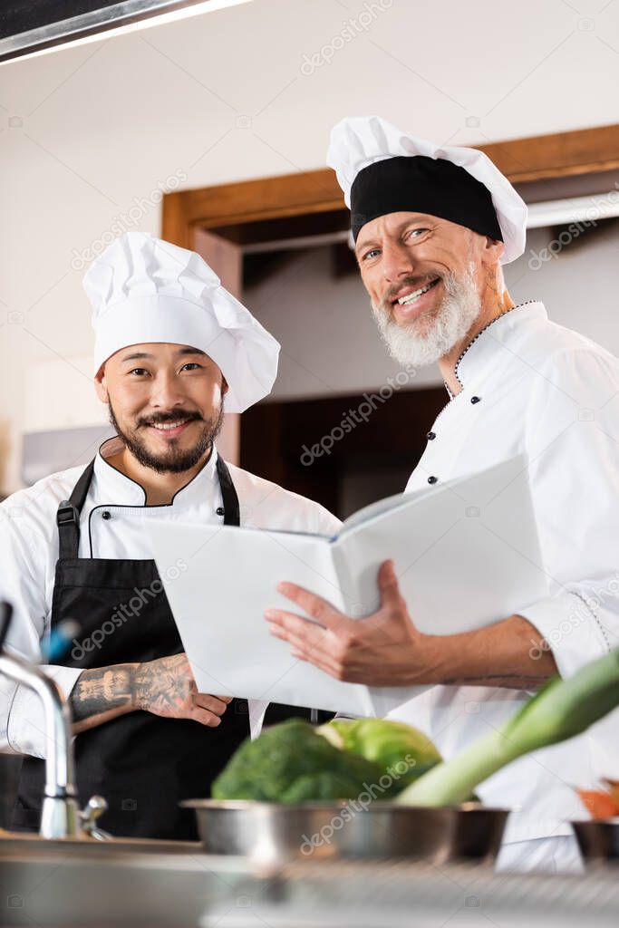 Mature chef holding cookbook near smiling asian colleague and blurred vegetables in kitchen 