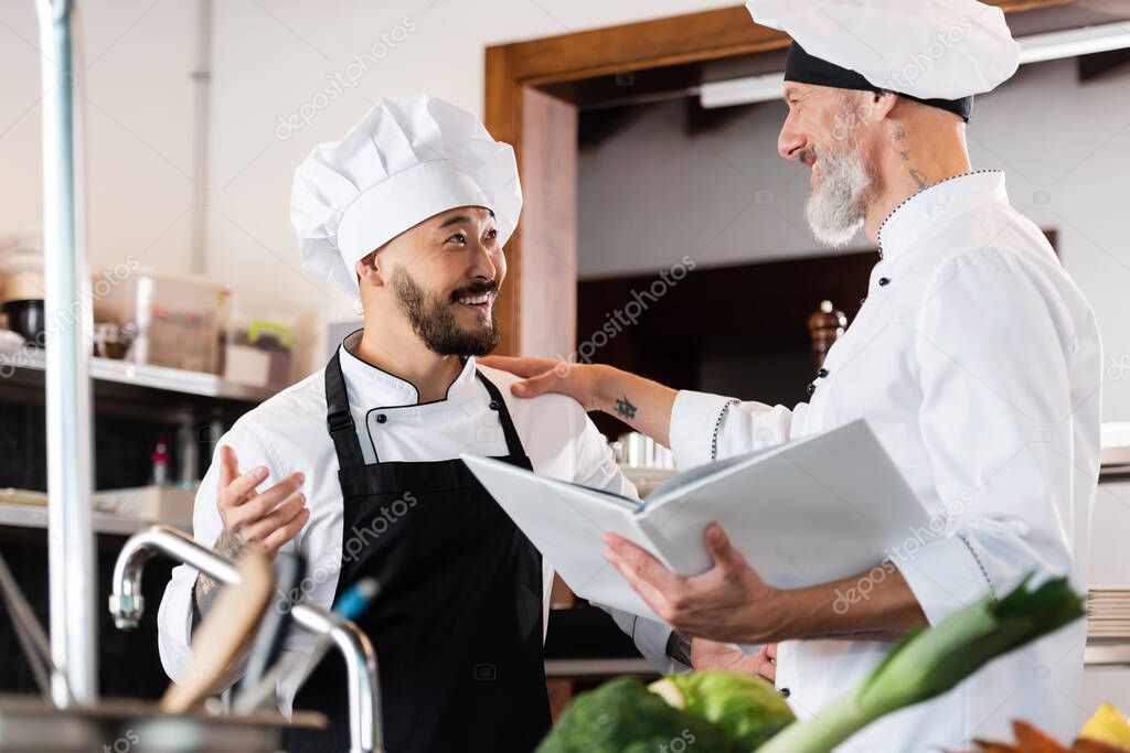 Positive chef holding cookbook while talking to asian colleague near faucet and vegetables in kitchen 