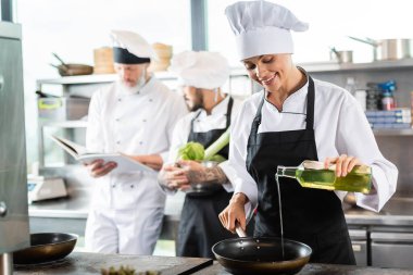 Önlüklü, gülümseyen bir aşçı, restoranın mutfağında yemek kitabı olan bulanık meslektaşlarının yanına zeytinyağı döküyor. 