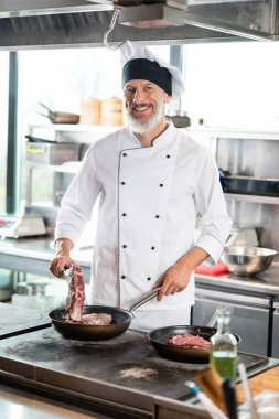 Üniformalı, gülümseyen bir aşçı restoran mutfağında et pişiriyor. 