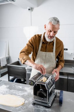 Olgun aşçı mutfakta makarna makinesine hamur koyuyor. 