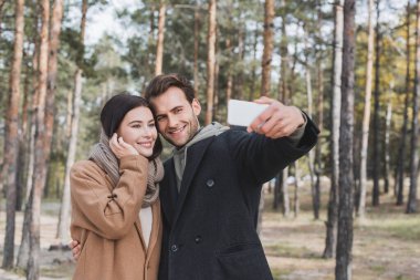 young man taking selfie with smiling girlfriend while walking in autumn park clipart