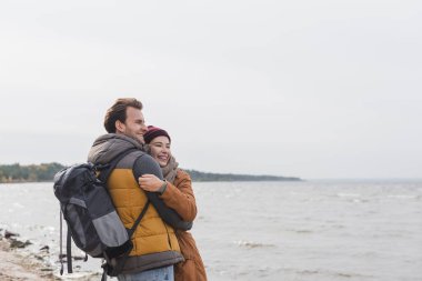Neşeli iki turist, denizin kenarında yürürken sarılıp bakışlarını kaçırıyorlar.