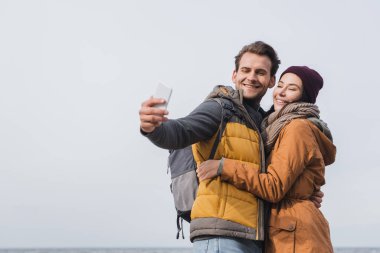 Mutlu yürüyüşçüler cep telefonlarıyla deniz kenarındaki mavi gökyüzüne selfie çekiyorlar.