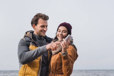 Nehir kenarında yürürken kız arkadaşının elinde akıllı telefonu gösteren gülümseyen adam.