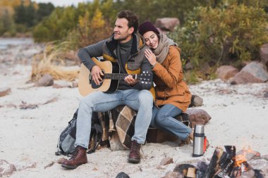 Genç adam yürürken şenlik ateşinin yanında gitar çalıyor.