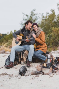 Şenlik ateşinin yanında otururken gitar çalan adama yaslanan mutlu adam.