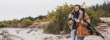 Gülümseyen çift, ekose battaniyeye sarılı, termo bardakları tutarken dışarı bakıyor, afiş