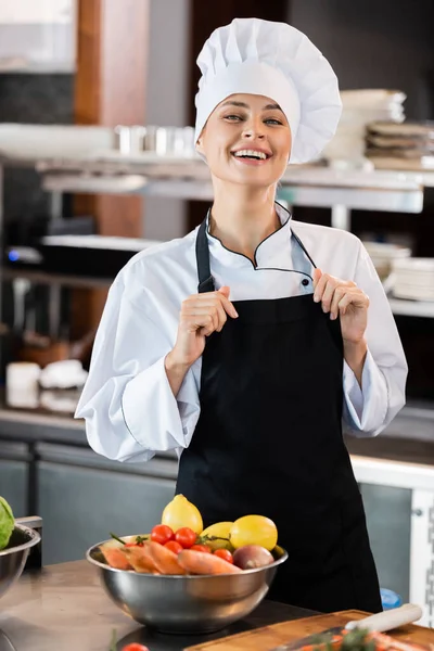 Positive Chef Touching Apron Looking Camera Fresh Vegetables Kitchen — Stock Photo, Image