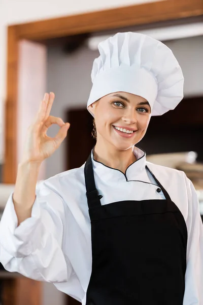 Positive chef in cap and apron showing okay gesture in kitchen