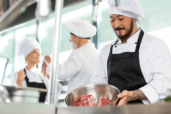 Sorrindo Asiático Chef Segurando Tigela Com Carne Perto Colegas Cozinha — Fotografia de Stock