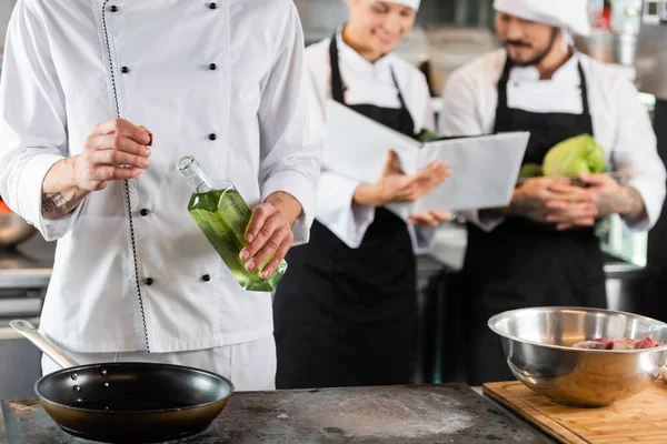 Szakács Gazdaság Üveg Olívaolaj Közelében Serpenyő Elmosódott Kollégák Szakácskönyv Konyhában — Stock Fotó