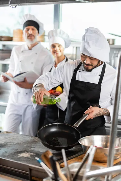 Asian Chef Pouring Oil Frying Pan Kitchenware Blurred Colleagues Cookbook — Stock Photo, Image