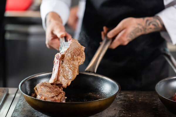 Cropped view of blurred chef preparing meat on frying pan in kitchen 