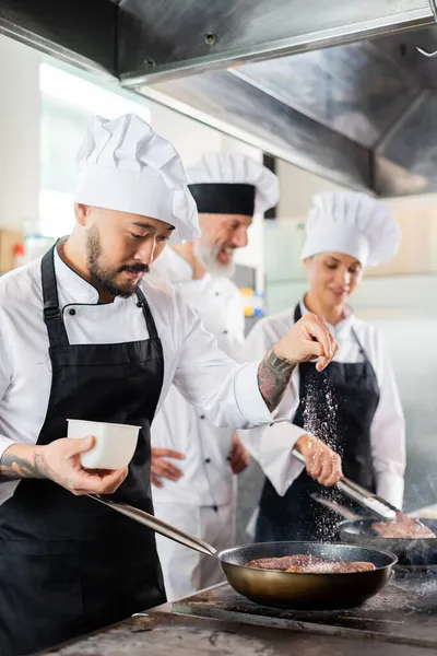 Asian Chef Pouring Salt Meat Cooktop Blurred Colleagues Kitchen — Stock Photo, Image