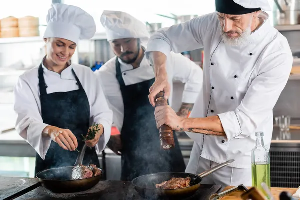 Chef Καρύκευμα Κρέας Στο Τηγάνι Κοντά Χαμογελαστοί Διαφυλετικοί Συνάδελφοι Δεντρολίβανο — Φωτογραφία Αρχείου