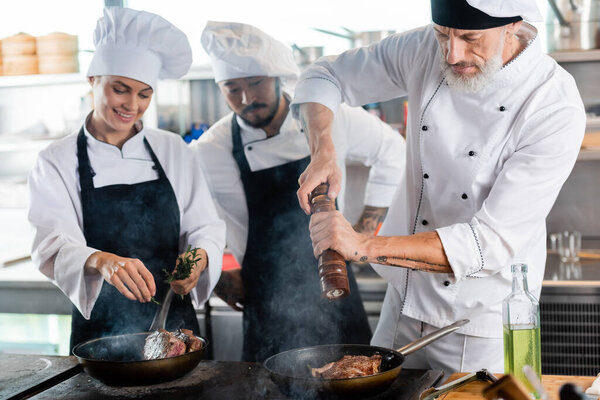 Chef seasoning meat on frying pan near smiling interracial colleagues with rosemary in kitchen 