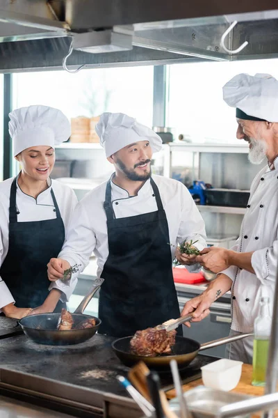 Chef Asiático Segurando Alecrim Perto Colegas Sorridentes Cozinhar Carne Cozinha — Fotografia de Stock