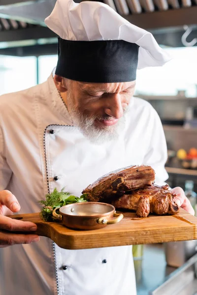 Middle aged chef in uniform and cap smelling roasted meat on cutting board in kitchen