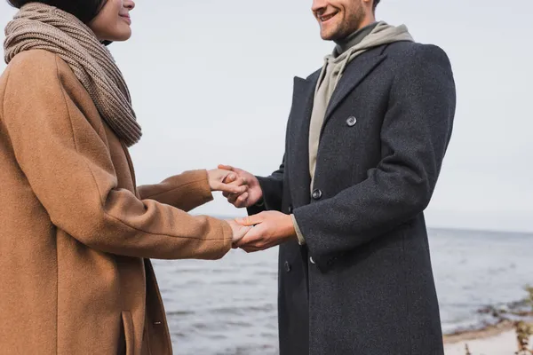 海の近くを歩いている間に手を取り合っている秋のコートで笑顔のカップルの眺め — ストック写真