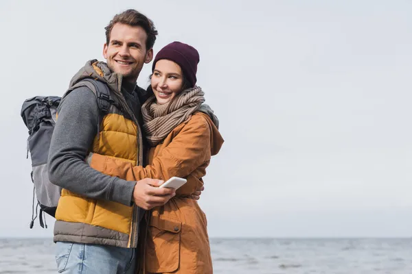 穿着秋装的快乐男人 一边拿着智能手机 一边在海边和女朋友拥抱 — 图库照片