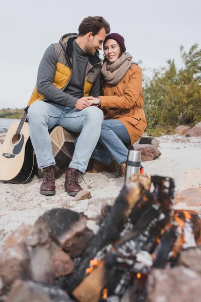 happy couple in autumn outfit holding hands while sitting near blurred bonfire