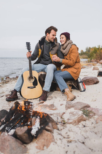 happy woman and man with guitar sitting near blurred bonfire at sea coast