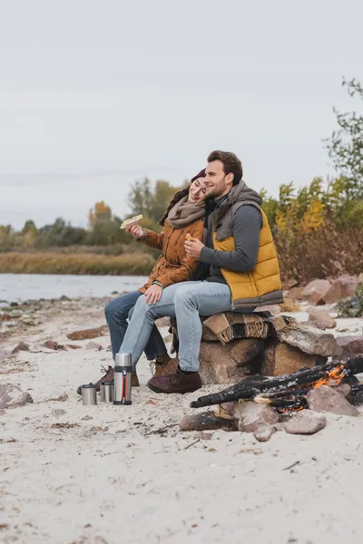 幸福的夫妇 带着三明治坐在热水瓶和篝火边的石头和毛毯上 — 图库照片