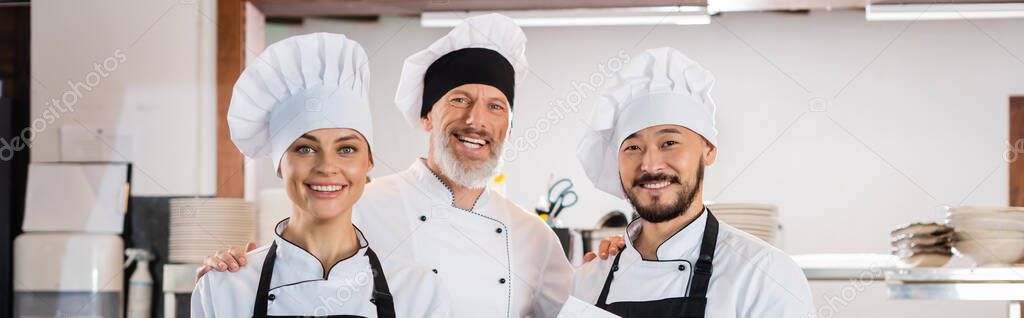 Smiling chef hugging interracial colleagues in caps in kitchen, banner 
