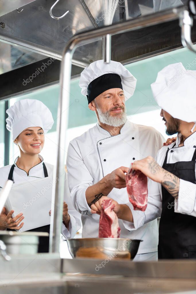 Chef pointing at raw meat near colleagues with cookbook in kitchen 