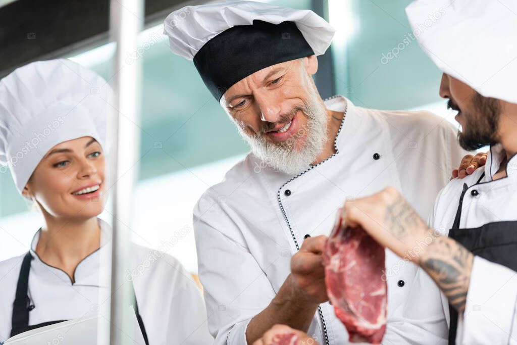 Smiling chef looking at blurred meat near colleagues with cookbook in restaurant kitchen 