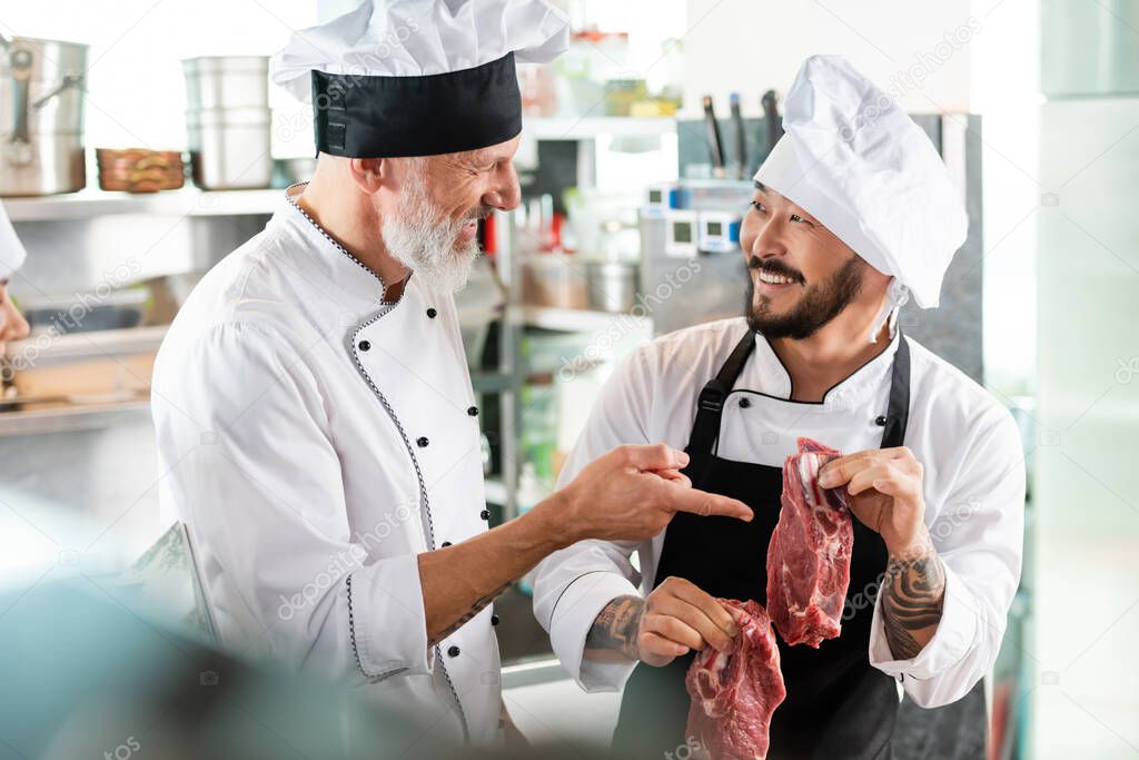 Smiling chef pointing at raw meat near tattooed asian colleague in kitchen 