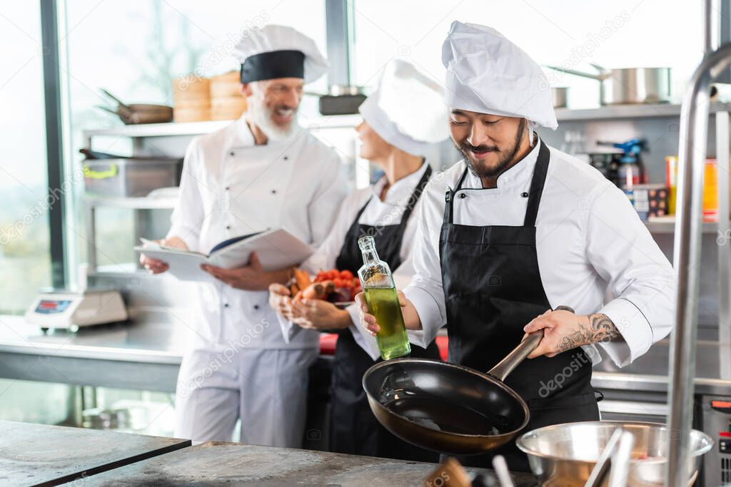 Smiling asian chef holding frying pan and olive oil near cooktop and blurred colleagues in kitchen 