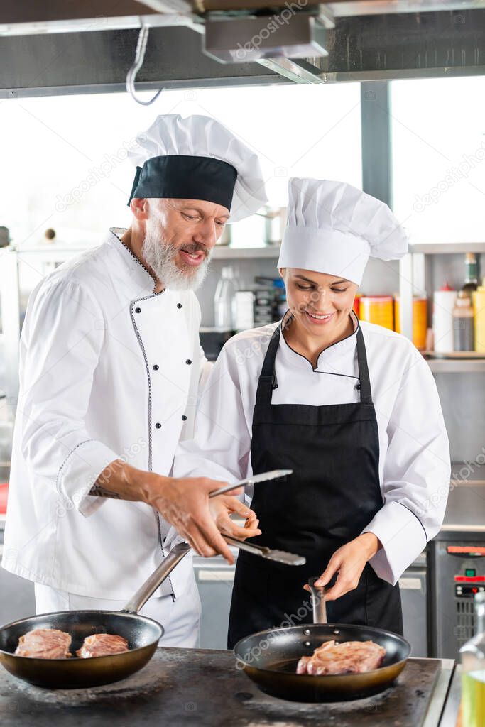 Chef holding tongs near smiling colleague and meat on frying pans in kitchen 