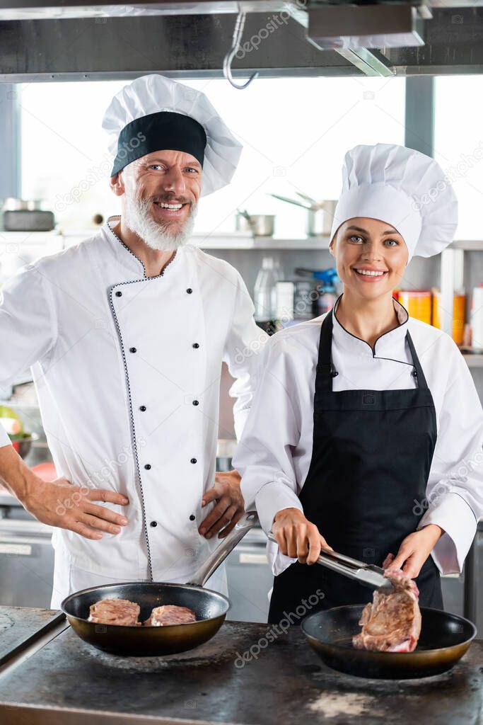 Smiling chefs in uniform looking at camera while cooking meat in kitchen 