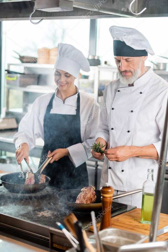Chef holding rosemary near smiling colleague roasting meat in kitchen 
