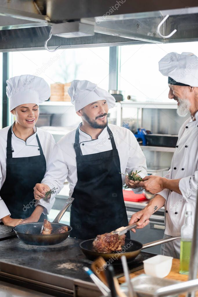 Asian chef holding rosemary near smiling colleagues cooking meat in kitchen 